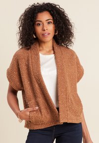 Frau mit lockigem Haar trägt goldene Creolen, ein weißes Oberteil und eine kurzärmlige, strukturierte braune Strickjacke mit Taschen und posiert vor einem beigen Hintergrund.