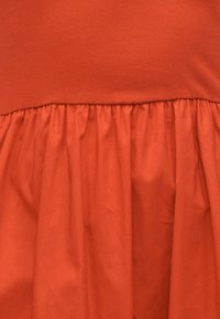 Ραφή από κόκκινο συλλεγμένο ύφασμα που συνδέει το λείο επάνω τμήμα με το πιο φαρδύ κάτω τμήμα, και τα δύο σε ζωντανό πορτοκαλί χρώμα.