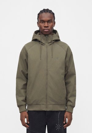 Jeune homme aux cheveux tressés portant une veste à capuche zippée verte et un pantalon noir clouté de métal sur un fond uni.