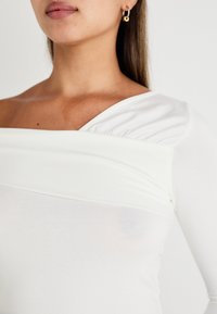 Žena v bílé topu s odkrytými rameny a dlouhými rukávy, na blízku vidíme její krk, rameno a zlaté kruhové náušnice.