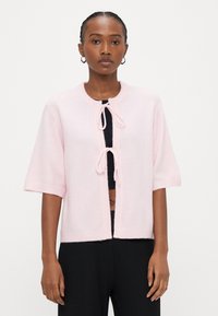 Light pink knitted cardigan with cropped sleeves, a round neckline, and a front tie closure, featuring soft texture and ribbed trim.