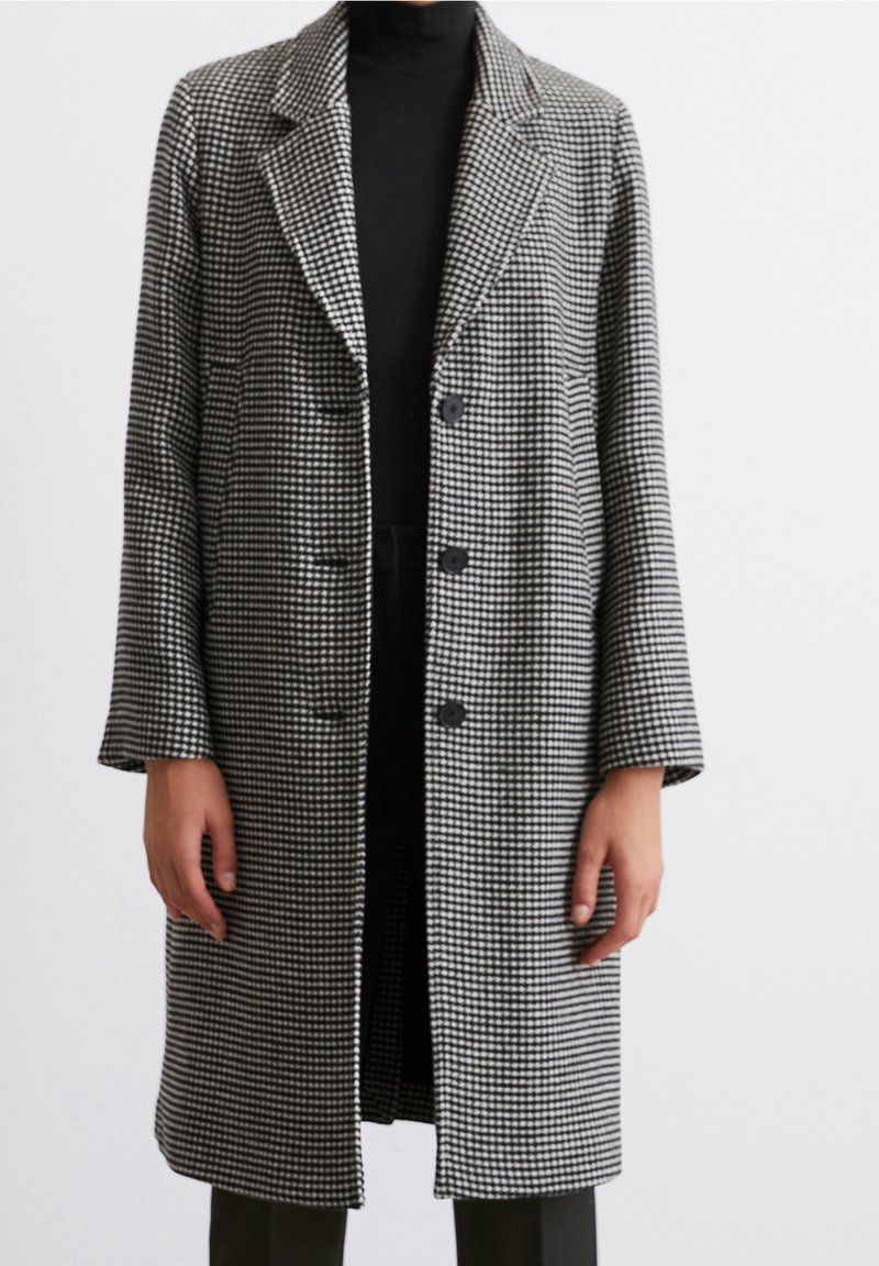 Personne portant un manteau à motif pied-de-poule noir et blanc longueur genou avec trois boutons, par-dessus un col roulé noir et un pantalon.