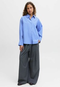 Camisa de botones a cuadros en azul claro con un corte relajado, combinada con pantalones grises de pierna ancha, que incluyen un bolsillo en la parte frontal y mangas largas.