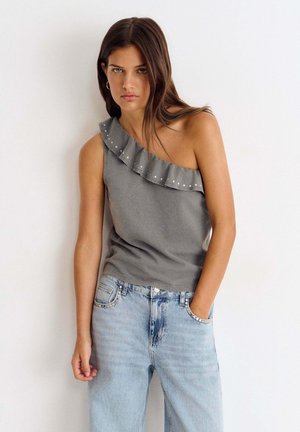 Mujer con cabello largo y castaño que lleva un top gris de un solo hombro con volantes y unos vaqueros azul claro de talle alto con tachuelas en los bolsillos.