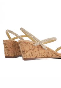 Kurkwig-sandalen met metallic gekruiste banden, versierd met kleine rhinestones. Beige kleur met een gestructureerde natuurlijke kurk.