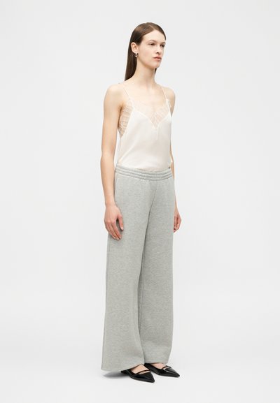 Femme debout portant un caraco blanc orné de dentelle, un pantalon de survêtement gris ample et des ballerines noires à bouts pointus sur un fond blanc.