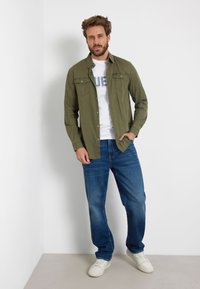 Tamsiai žalia mygtukų marškiniai su dviem krūtinės kišenėmis, po jais balta marškinė su grafika, mėlynos džinsinės kelnės ir baltos sportinės avalynės.