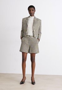 Blazer och shorts-set i houndstooth-mönster i kräm, svart och olivgrön. Blazern har en figurnära passform; shortsen har en avslappnad skärning. Svarta pumps.