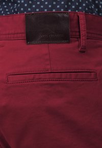 Röda bomullshängselbyxor med en brun läderetikett märkta "BOSS ORANGE." Har en bakficka med subtila sömnadsdetaljer.