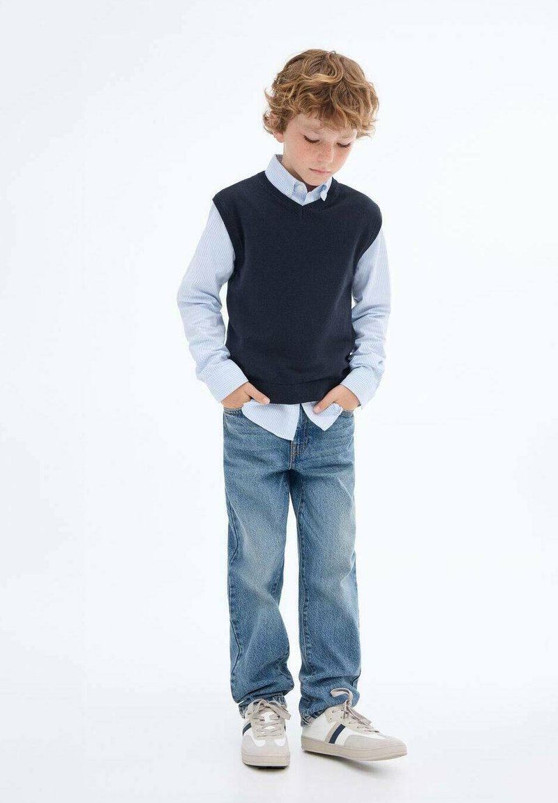 Námořnický bezrukávový svetr přes světle modrou košili s knoflíky, doplněný modrými džínami a béžovými teniskami s tmavými pruhy.