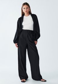 Zwarte longsleeve shirt, witte geribde top, hoge zwarte wijde broek en zwarte instap-sandalen met gouden accenten.