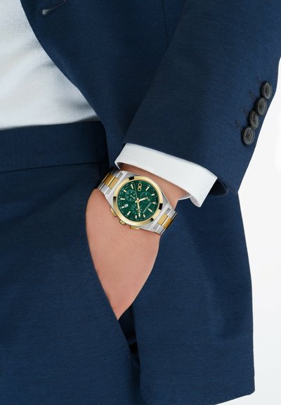 Reloj de plata y oro con una esfera verde, exhibido en una muñeca, que cuenta con una caja cuadrada, funciones de cronógrafo y una pulsera de metal pulido.