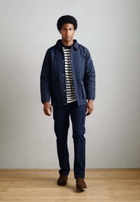 Marineblå quilted jakke med to frontlommer over en sort og hvid stribet skjorte. Iført mørke bukser og brune støvler, stående på trægulv.