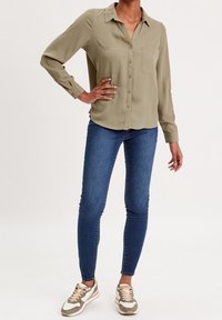 Chemise boutonnée vert olive avec deux poches avant, associée à un jean skinny bleu et des baskets marron clair avec des accents blancs et un design texturé.