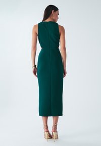 Temno zelena midi obleka brez rokavov s prilegajočim se krojem, rahlo zadrgo na hrbtu in izrezanimi detajli na straneh. Stilizirana z bež sandali s trakovi.