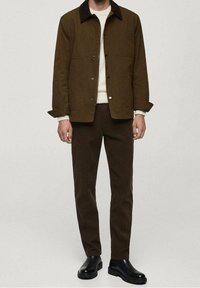 Bruine jas met een donkere kraag, knoopsluiting en twee voorzakken, gecombineerd met een bruine broek en zwarte enkellaarsjes.