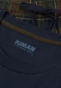Mörkblå T-shirt med rund halsringning och grön kant, med "IUMAN"-logotypen i ljusgrått. Under finns en rutigt tyg i grönt, rött och gult.