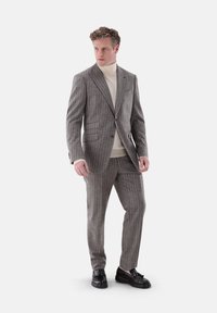 Grauer Nadelstreifenanzug mit slim fit, strukturiertem Stoff, Reverskragen und zwei Fronttaschen, kombiniert mit einem cremefarbenen Rollkragenpullover und schwarzen Schuhen.