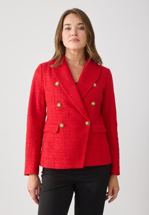 Chaqueta roja texturizada con diseño de doble botonadura, botones dorados y bolsillos frontales. Corte ajustado con solapas anchas. Combinada con pantalones negros.
