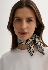 Czarnobiała jedwabna chusta w wzory, związana wokół szyi, z motywami paisley i kropeczkami, noszona na gładkiej białej koszuli.