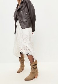 Giacca da motociclista in pelle marrone, vestito bianco in pizzo, stivaletti beige slouchy con punta appuntita e tacco in legno. Orlo dettagliato in pizzo sul vestito.