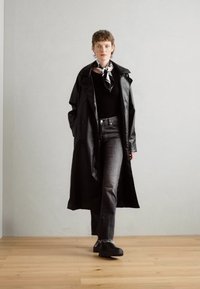 Black, knee-length raincoat with large buttons, worn over a black top and gray jeans. Complemented by a patterned scarf and black shoes.