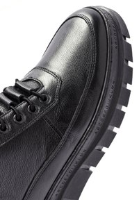 Zapato de cuero negro con parte superior texturizada, puntera redonda y suela de goma gruesa con tacos pronunciados y patrón de alfabetos en relieve.