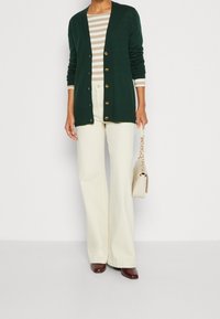 Cárdigan verde oscuro con botones dorados sobre un top a rayas crema y beige, combinado con pantalones beige de pierna ancha y zapatos granate, sosteniendo una bolsa beige.