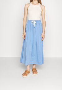 Gonna maxi blu chiaro con cintura elastica e cordoncino bianco. Abbinata a una canotta bianca e sandali marroni con suola spessa.