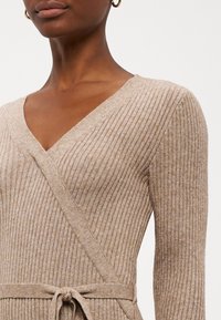Pull en maille côtelée beige avec col en V et manches longues, doté d'un design cache-cœur et d'une ceinture à nouer à la taille. Texture douce et coupe élégante.