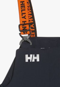 Marinblå overaller har orange justerbara hängslen med svart varumärke, en silverfärgad logo på bröstet och en kraftig metallspänne.