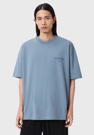 Jeune personne aux longs cheveux foncés portant un T-shirt surdimensionné AllSaints bleu clair et un pantalon noir, posant devant un fond uni.