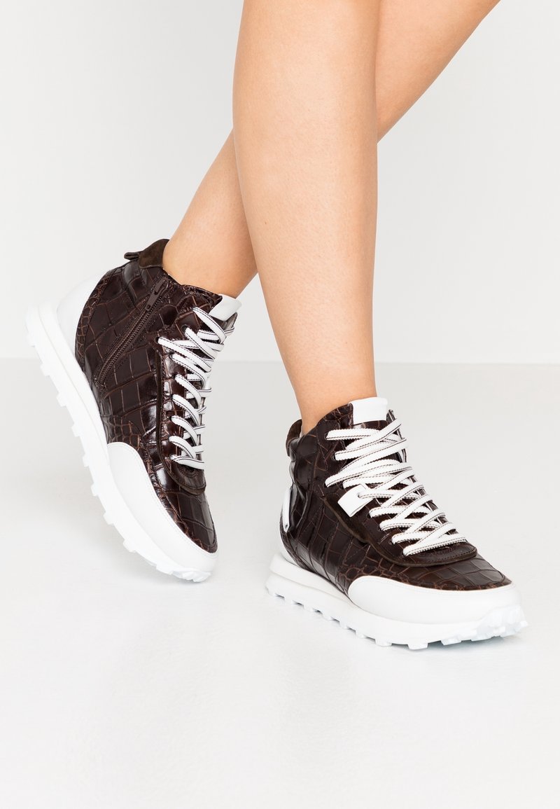 Jambes portant des baskets montantes en cuir crocodile marron foncé avec des semelles et des lacets blancs sur une surface blanche.