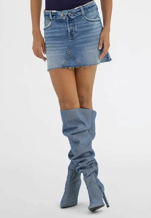 Donna che indossa una mini gonna di denim azzurro chiaro e stivali alti sopra il ginocchio abbinati, con tacco, in posizione eretta davanti a uno sfondo semplice.