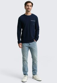 Navyblaues Langarmshirt aus strukturiertem Stoff, mit einer weißen Taschenakzent. Kombiniert mit hellblauen Jeans und weißen Turnschuhen.