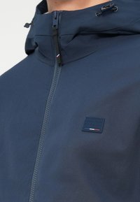 Mornarsko modra jakna z visokim ovratnikom, sprednjim zadrgo in logotipom "JACK & JONES PREMIUM" v temno modri barvi s rdečimi in belimi detajli.