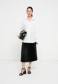 Biała koszula z guzikami, czarna plisowana spódnica i czarne sandały na obcasie z paskami. Modelka trzyma czarną torebkę typu clutch o teksturowanej powierzchni z zaokrąglonym kształtem.