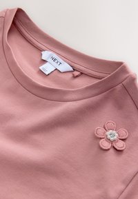 Roza majica z okroglim izrezom, z okrasnim vezenim cvetom s perlami na levi strani. Iz mehke bombažne tkanine.