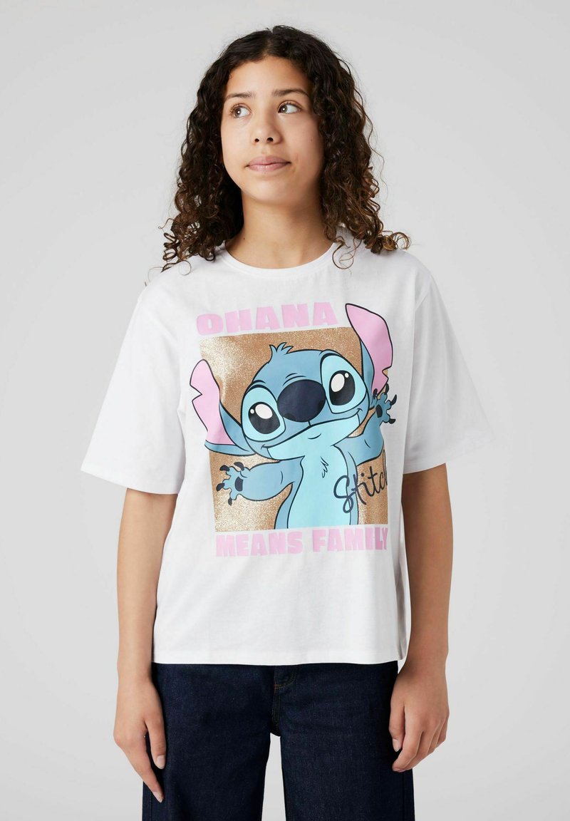 Jongere met krullend haar en een wit T-shirt met een Stitch-tekening en de tekst "Ohana Means Family" tegen een effen achtergrond.