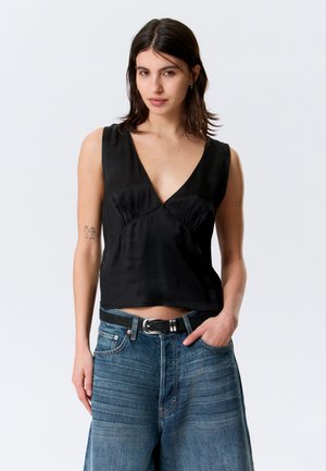 Mujer con cabello oscuro hasta los hombros, lleva un top negro sin mangas con escote en V y jeans azules de talle alto con cinturón negro, de pie con una mano en el bolsillo.