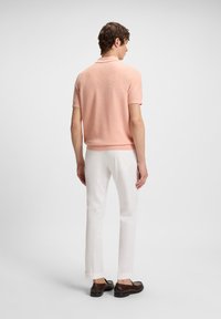 Homem em posição de costas, vestido com uma camisa polo de manga curta em pêssego, calças brancas e sapatos mocassim castanho-escuros, contra um fundo claro e uniforme.