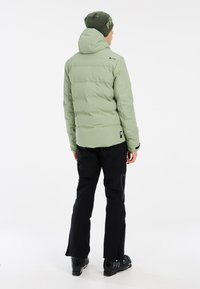 Hellgrüne Pufferjacke mit Kapuze, mit Reißverschlüssen versehenen Taschen und schwarzen Skihosen. Das Outfit wird mit schwarzen Skischuhen kombiniert. Einfaches, funktionales Design.