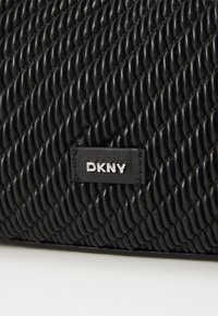 Μαύρη τσάντα χεριού DKNY με υφή, πλεγμένο σχέδιο και γυαλιστερό φινίρισμα. Η ετικέτα με το λογότυπο εμφανίζεται prominently σε ασημί στο πλάι.