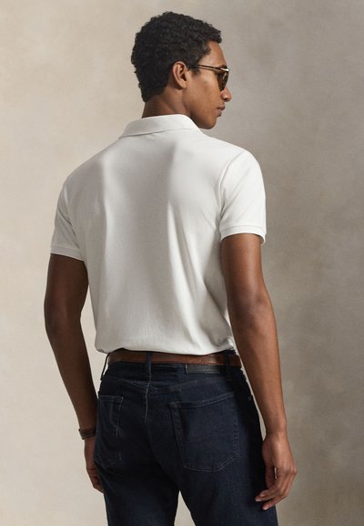 Rövid göndör hajú férfi napszemüvegben, fehér rövid ujjú galléros ingben, sötét farmerban és barna övvel, semleges háttér előtt állva.