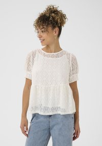 Haut en dentelle blanche avec de courtes manches ballons, présentant un motif de diamants et un ourlet peplum, associé à un jean en denim bleu clair.