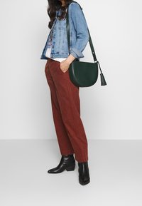 Džínová bunda, bílý grafický tričko, hnědé kordové kalhoty, černé kotníkové boty, zelená kožená taška přes rameno s třásněmi.