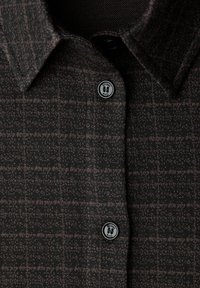 Camicia a quadri scura con tessuto strutturato. Presenta un colletto e due bottoni neri con logo. Il motivo è costituito da quadrati marrone chiaro su base scura.