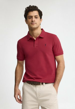 Man draagt een rood polohemd met korte mouwen en beige broek met een bruine riem, staand met één hand in de zak.