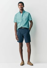 Hellblaue kurzärmlige Button-Down-Hemd, marineblaue Shorts, schwarzer Gürtel, beige Slipper; glattes Material, lässige Passform, minimalistischer Stil, keine Muster.