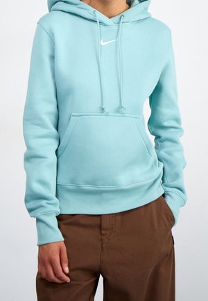 Person iført en lyseblå Nike-hoodie med frontlomme og brune bukser, med hænder delvist i lommerne mod en ensfarvet baggrund.